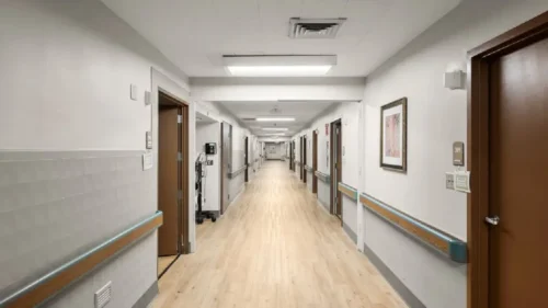 A long, empty hallway with wood floors, handrails, and closed doors on both sides, with a framed picture on the right wall