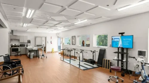 A physical therapy room with parallel bars, exercise equipment, a kitchenette, and two wheelchairs