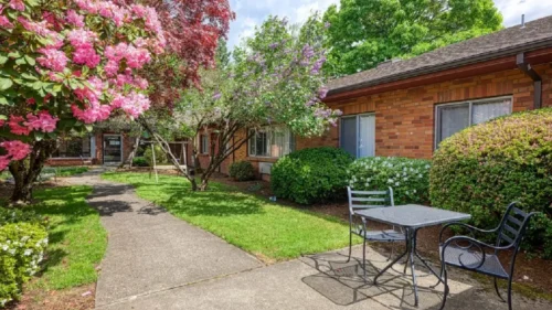 An outdoor patio area with a table and chairs next to a walkway, surrounded by lush green grass, a brick building, and blooming trees
